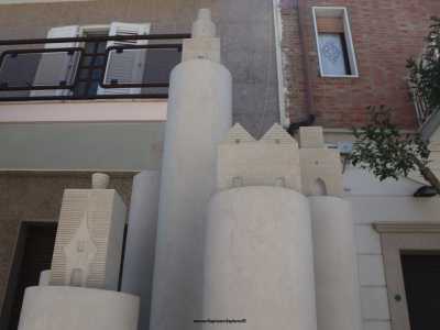 La base scalpellata simboleggia l'estrazione della roccia "grezza", mentre il corpo centrale levigato richiama la maestria degli artigiani. In cima a ciascun fusto si stagliano infine palazzi in miniatura di varia forma a righe orizzontali...